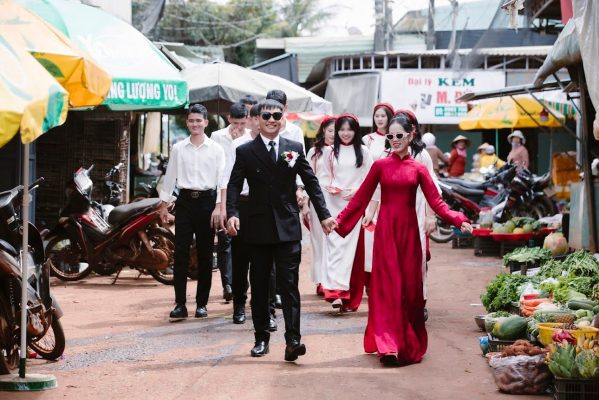 Chú rể mặc áo dài và vest nắm tay nhau đi giữa khu chợ truyền thống